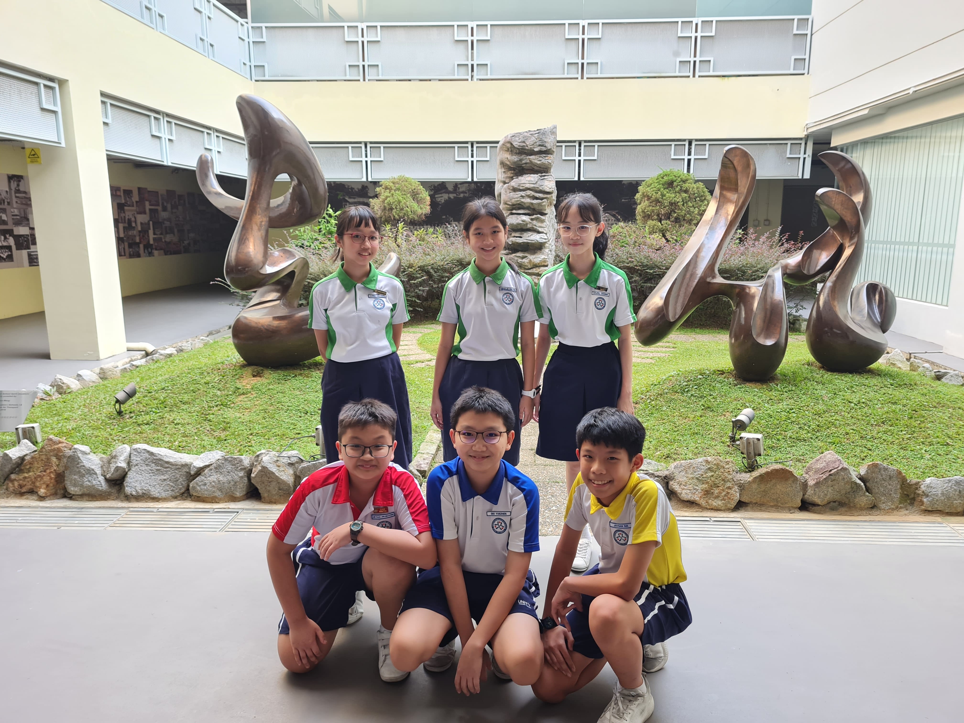 Six school children in uniforms pose in two rows before a garden courtyard with large bronze sculptures.